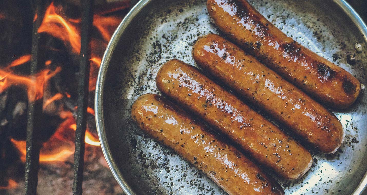 Four hot dogs grilling in a pan over open flame, by Rachel Clark via Unsplash
